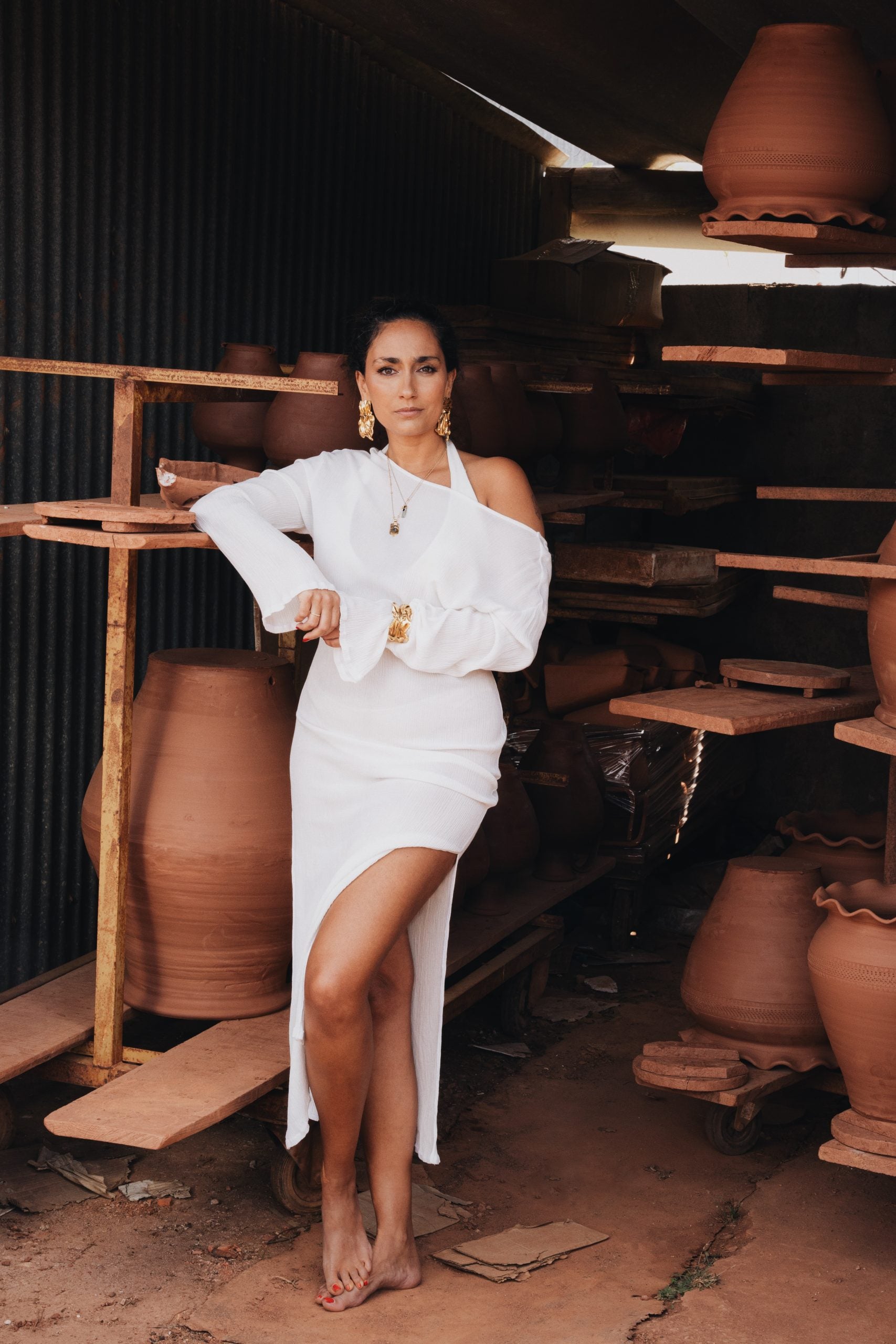 Woman in a white off-shoulder dress with gold jewelry standing barefoot among clay pottery.