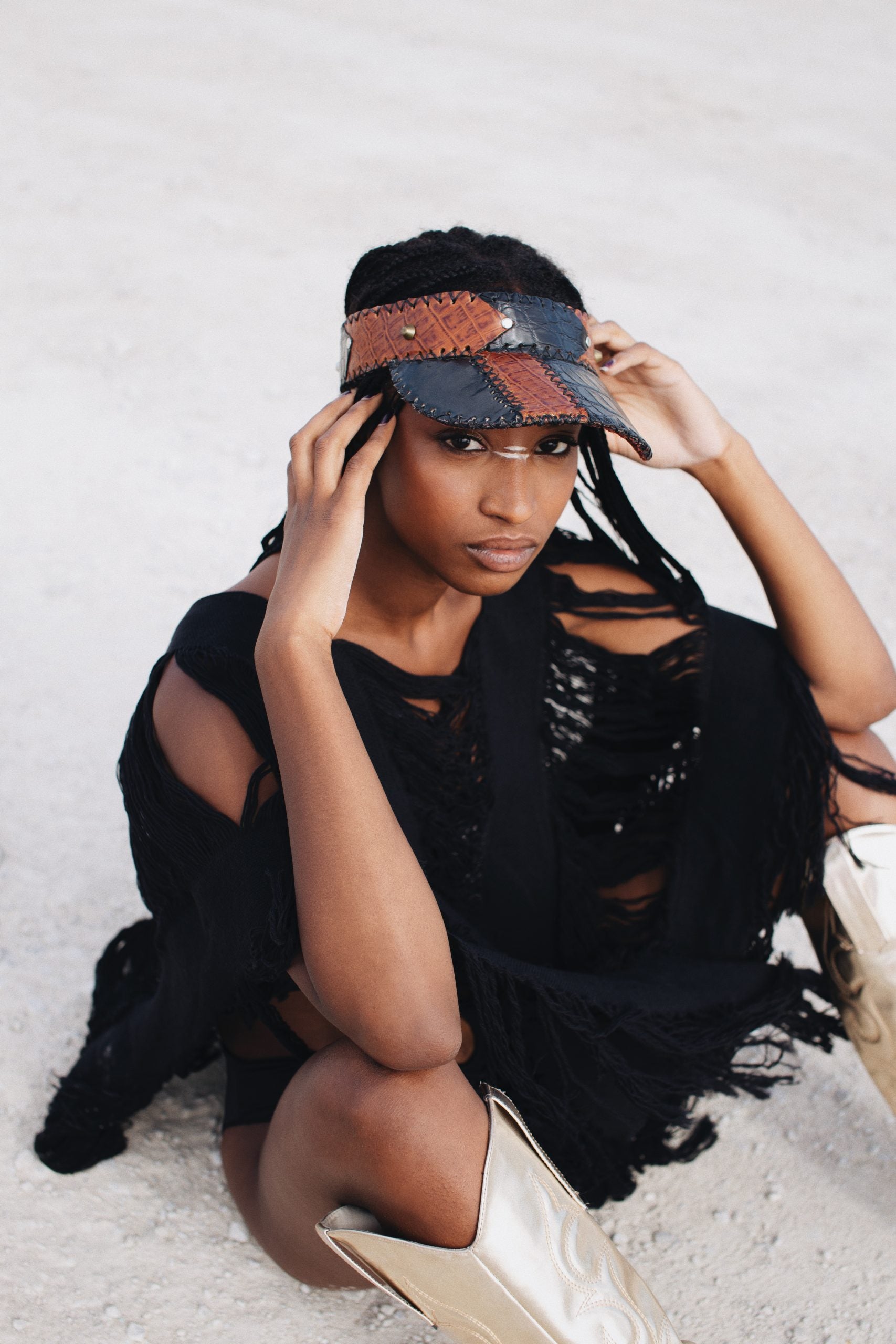 Woman in handmade patchwork visor, distressed black poncho, and metallic boots on sand