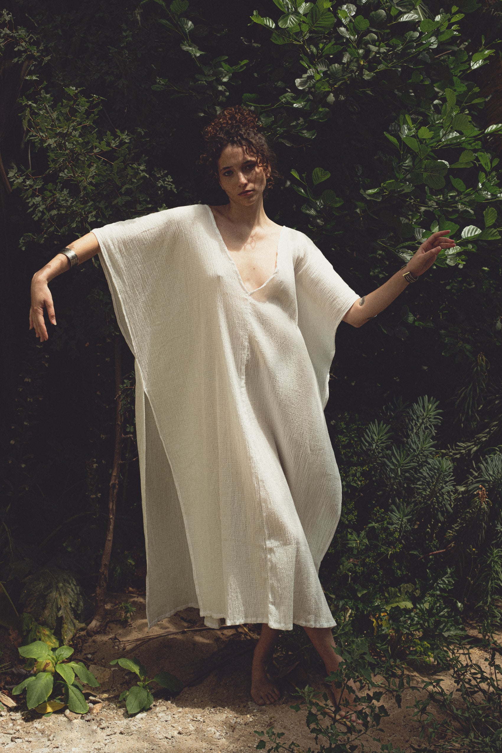 Woman in a loose white handmade dress standing barefoot in a lush green garden