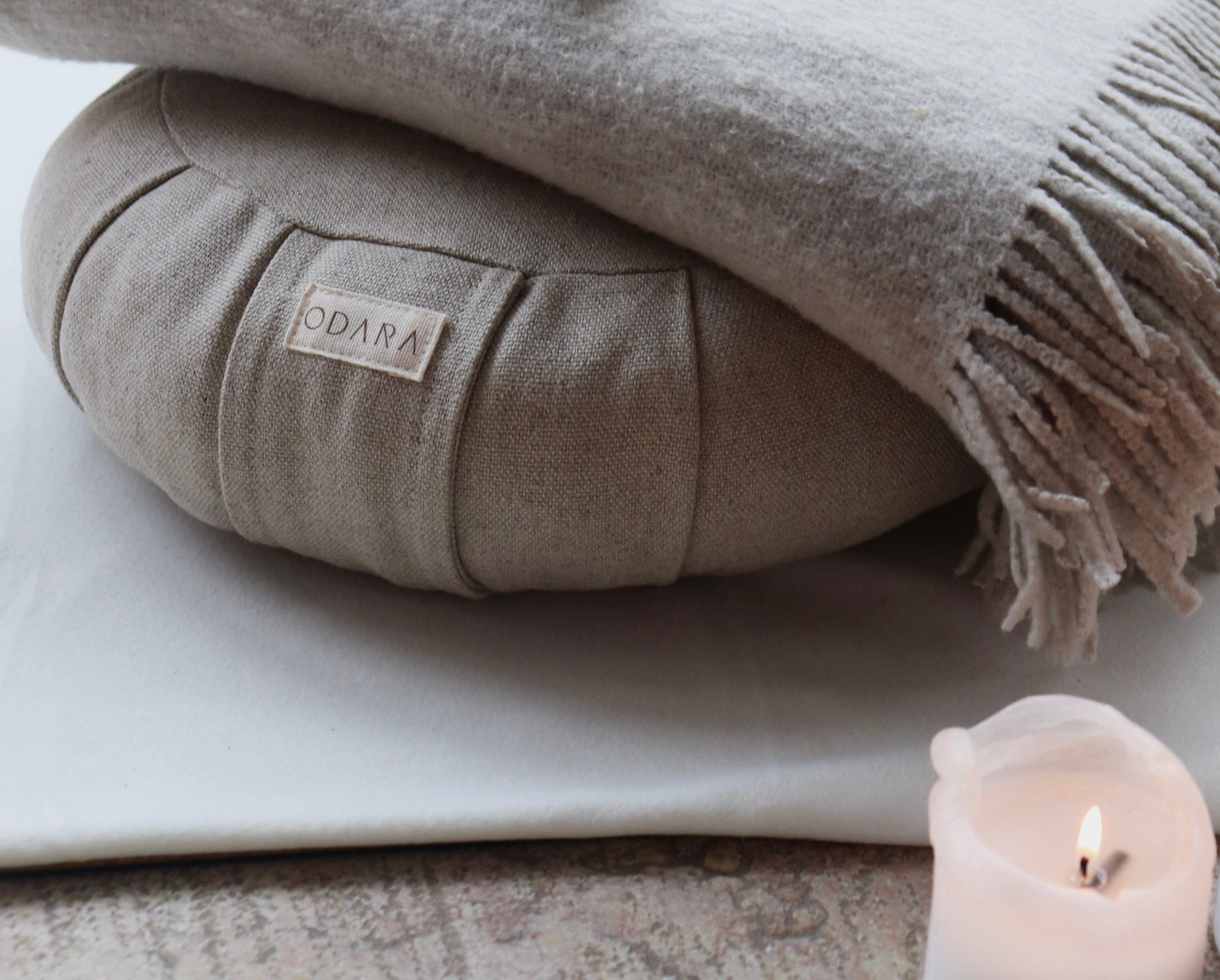 Neutral linen meditation cushion, fringed throw, and burning candle on soft surface