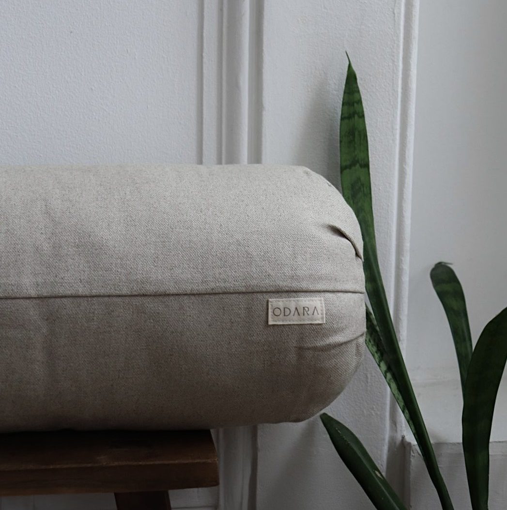 Beige handmade bolster pillow with fabric label on wooden bench beside green plant